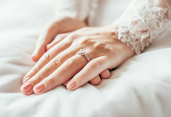 Hand in a lace sleeve wearing a diamond engagement ring, resting on white fabric